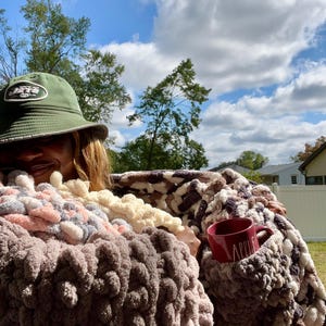 May include: A person is wrapped in a multi-colored chunky knit blanket with a red mug that says "Apple" on it. The person is wearing a green baseball cap with the word "Jets" on it.