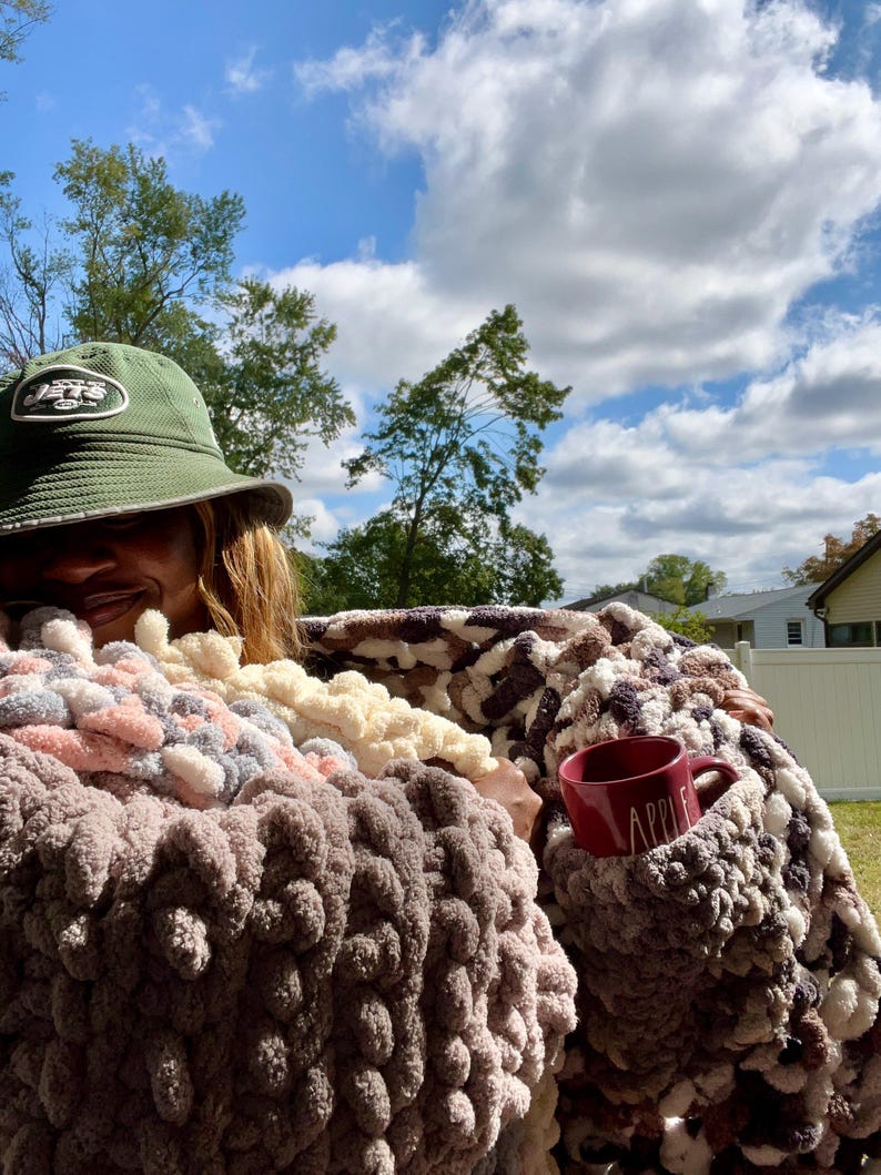 May include: A person is wrapped in a multi-colored chunky knit blanket with a red mug that says "Apple" on it. The person is wearing a green baseball cap with the word "Jets" on it.
