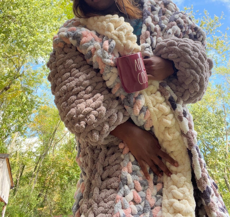 May include: A chunky knit cardigan with a cream and pink color scheme. The cardigan is open in the front and has a relaxed fit. The person wearing the cardigan is holding a red mug that says "Apple Cider".