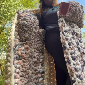 May include: A long, chunky knit cardigan with a cream border and a multi-colored yarn body. The yarn is a mix of pink, gray, and white. The cardigan is open in the front and has a large pocket on the left side.