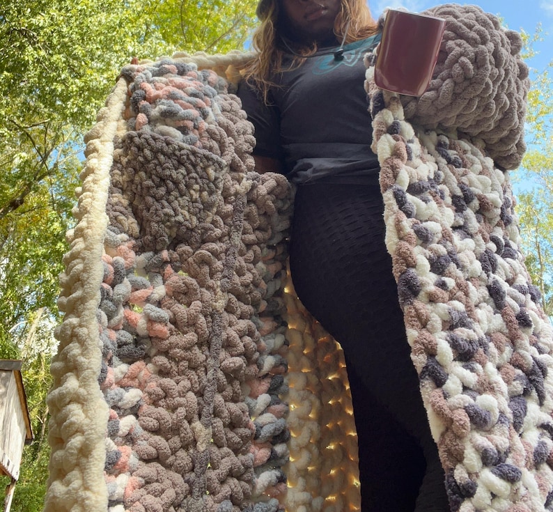May include: A long, chunky knit cardigan with a cream border and a multi-colored yarn body. The yarn is a mix of pink, gray, and white. The cardigan is open in the front and has a large pocket on the left side.