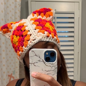 Könnte beinhalten: Eine weiße Häkelmütze mit einem bunten Granny-Square-Muster in den Farben Pink, Orange und Gelb.