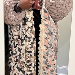 May include: A person wearing a long, multi-colored crocheted cardigan with a pink and gray pattern. The cardigan has large, chunky stitches. The person is also wearing a green bucket hat with a white brim and a white watch. The person is holding a red mug with the text "CUDDLE" in white.