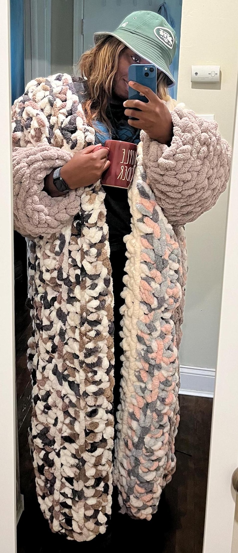 May include: A person wearing a long, multi-colored crocheted cardigan with a pink and gray pattern. The cardigan has large, chunky stitches. The person is also wearing a green bucket hat with a white brim and a white watch. The person is holding a red mug with the text "CUDDLE" in white.
