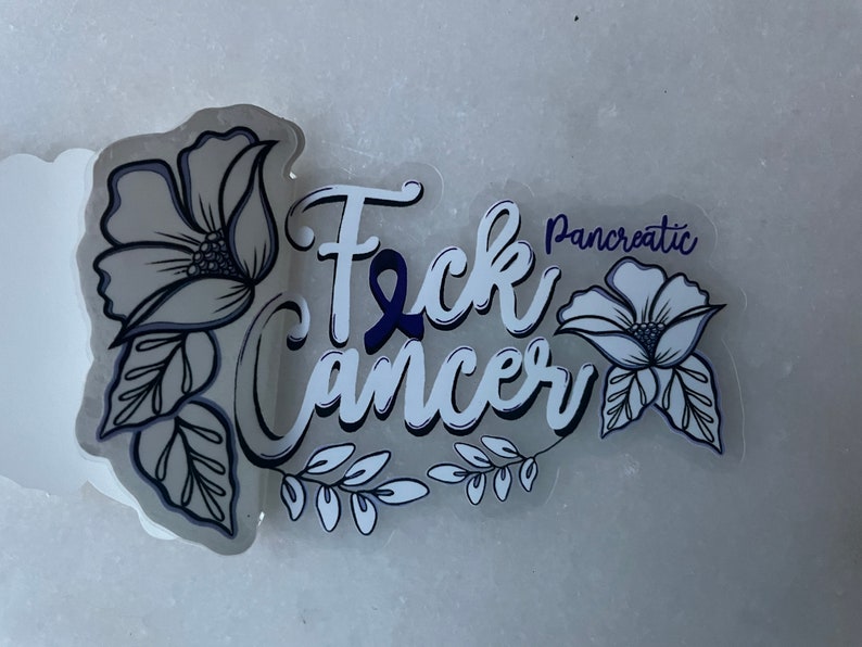 May include: White sticker with the text "F*ck Cancer" in a stylized font with a blue ribbon. The text "Pancreatic" is above the word "Cancer". The sticker is decorated with black and white floral designs.