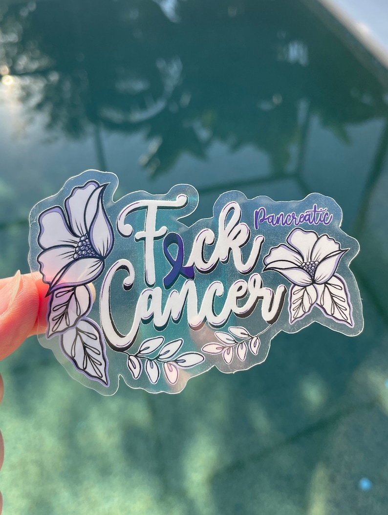 May include: A clear sticker with the words "F*ck Cancer" in white with a purple ribbon and the word "Pancreatic" in smaller white letters. The sticker is decorated with white flowers and leaves.