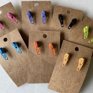 May include: Assortment of colorful zipper earrings displayed on brown card stock. The earrings are in various colors including pink, purple, blue, orange, yellow, black, and green.