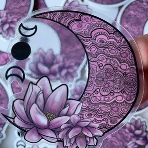 May include: A clear sticker with a crescent moon design. The moon is filled with a purple and black mandala pattern. Two purple flowers are at the bottom of the moon.