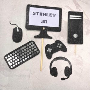 May include: A set of black glittery gaming-themed cake toppers including a computer monitor with the text STANLEY 30, a computer tower, a keyboard, a gaming mouse with a cord, a game controller, and a headset, all featuring wooden sticks.