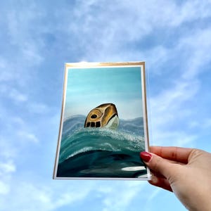 Puede incluir: Una tarjeta de felicitación con una ilustración artística de una tortuga marina en el océano. La obra de arte muestra la cabeza de una tortuga con un diseño detallado de caparazón, sobre agua y cielo azules. La tarjeta tiene un borde dorado.