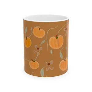 Puede incluir: Una taza de cerámica con fondo marrón sólido que presenta un patrón repetitivo de calabazas de color naranja claro, pequeñas hojas verdes e ilustraciones de flores de color marrón claro. El interior es blanco.