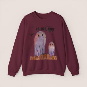 Puede incluir: Una sudadera de manga larga color granate con un estampado gráfico de dos fantasmas en un huerto de calabazas y el texto FA-BOO-LOUS encima. Los fantasmas están dibujados al estilo de lápices de colores con pequeños murciélagos negros.