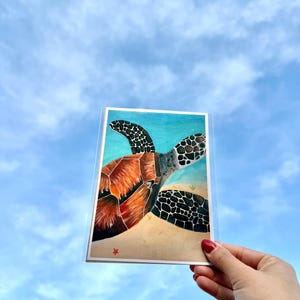 Puede incluir: Una pintura a la acuarela de una tortuga marina con un caparazón marrón y aletas negras y blancas. La tortuga está representada en una playa arenosa con un fondo azul océano. La obra de arte se sostiene contra un cielo azul.