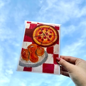 Op de afbeelding: Een wenskaart met een schilderij van een pizza op een houten pizzaschep en gefrituurd voedsel op een bord, tegen een rood-witte geruite achtergrond. De kaart wordt tegen een blauwe lucht gehouden.
