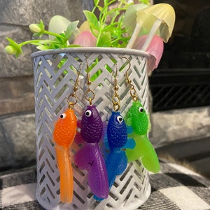 May include: Four pairs of dangle earrings featuring colorful fish charms. The fish are orange, purple, blue, and green. The earrings are hanging from a white wire rack.