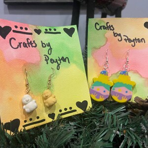 May include: Two pairs of handmade earrings on display cards. The first pair features a white snowman charm and the second pair features a yellow and green snowman charm. Both cards have the text "Crafts by Payton" written on them.