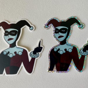 May include: Two stickers of Harley Quinn, a comic book character, giving the middle finger. One sticker is a standard print, the other is a holographic sticker.