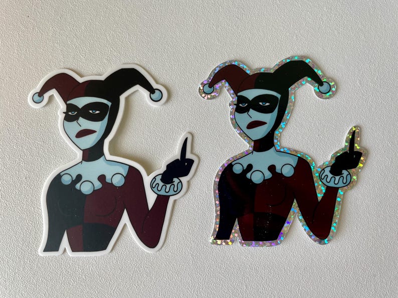 May include: Two stickers of Harley Quinn, a comic book character, giving the middle finger. One sticker is a standard print, the other is a holographic sticker.