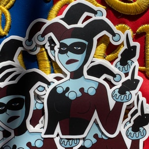 May include: A sticker featuring a cartoon character with black and red jester's clothing, a black mask, and blue skin. The character is giving a middle finger.