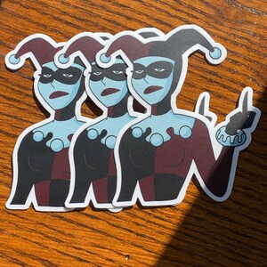 May include: Three stickers of a cartoon character with blue skin, red and black jester's clothing, and a black mask. The character is giving a middle finger.