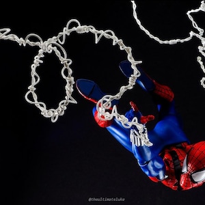 Puede incluir: Una figura de acción de Spider-Man roja y azul colgada de líneas de telaraña blancas sobre un fondo negro. La figura está en una pose dinámica con una pierna extendida y la otra doblada. Las líneas de telaraña están enredadas y crean una sensación de movimiento.