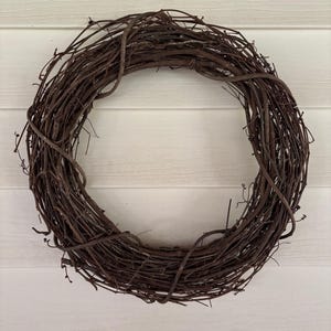 May include: A circular wreath made of intertwined brown twigs. The wreath is set against a white, horizontally-paneled background. The twigs are a natural brown color, creating a rustic and organic aesthetic.