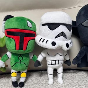 May include: Four plush Star Wars character dolls: a green alien, Boba Fett, a Stormtrooper, and Darth Vader. The dolls are soft and colourful, with detailed features and iconic designs. They are arranged in a row, perfect for fans of the franchise.