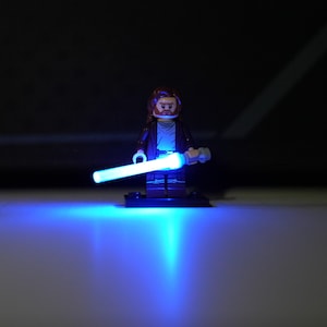 May include: A LEGO minifigure with brown hair and a beard, wearing a dark brown robe over a tan tunic, holding a bright, glowing blue lightsaber that casts a vibrant light onto the surface below, standing on a small black rectangular base.