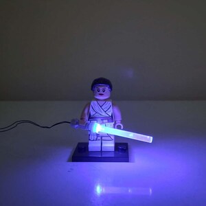 May include: A LEGO minifigure with dark hair and a white outfit standing on a black rectangular base. The figure holds a light-up blue plastic lightsaber accessory that emits a bright glow, connected by a thin black wire extending to the left side.