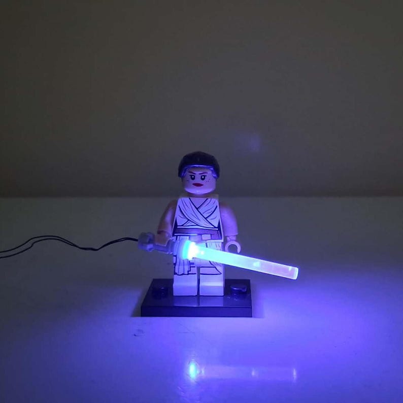 May include: A LEGO minifigure with dark hair and a white outfit standing on a black rectangular base. The figure holds a light-up blue plastic lightsaber accessory that emits a bright glow, connected by a thin black wire extending to the left side.