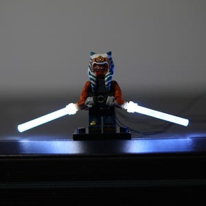 May include: A Lego minifigure of a character with orange skin and blue and white headgear, standing on a black base. The figure holds two glowing white light-up plastic rods that extend outward from each hand, connected by thin black wires.