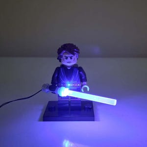 May include: A plastic minifigure with brown hair and a stern facial expression, wearing a dark tunic and pants. The figure stands on a black rectangular base and holds a translucent light blue lightsaber that is illuminated with a bright blue LED light.