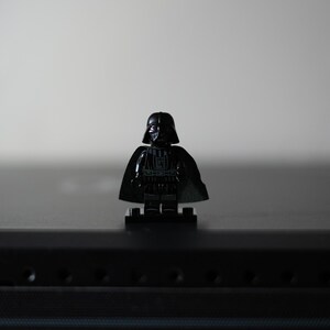 May include: A black plastic minifigure featuring a detailed helmet, a dark cape, and printed torso armor. The character stands on a small black rectangular base, showcasing a glossy finish on the helmet and chest plate with subtle gray accents.