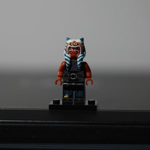 May include: A plastic minifigure featuring a character with orange skin, blue and white striped head-tails, and a gold headpiece. The figure wears a dark gray sleeveless tunic over a black outfit with a belt, standing on a black rectangular base.
