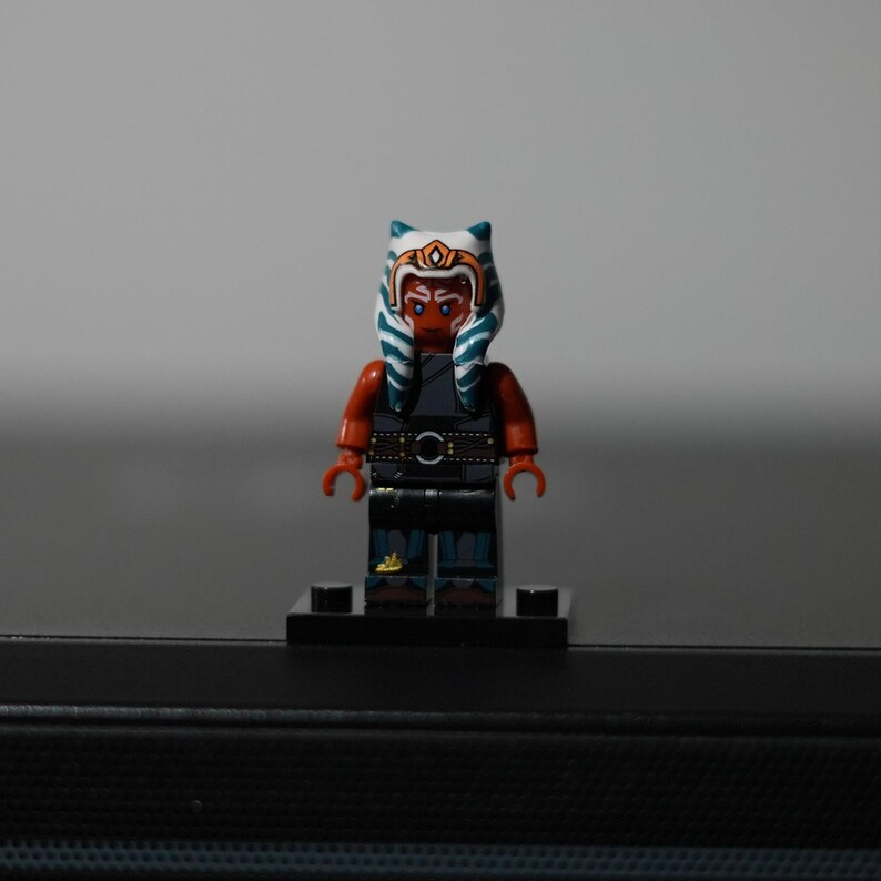 May include: A plastic minifigure featuring a character with orange skin, blue and white striped head-tails, and a gold headpiece. The figure wears a dark gray sleeveless tunic over a black outfit with a belt, standing on a black rectangular base.