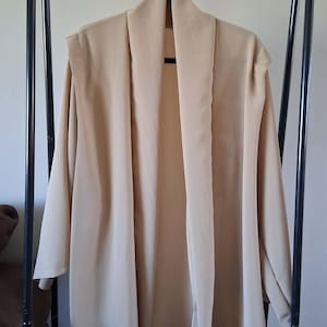 May include: A beige open-front cardigan with a wide shawl collar and three-quarter length sleeves. The garment features a smooth, solid-colored fabric with a relaxed, flowing silhouette that hangs straight down from the shoulders.