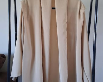 May include: A beige open-front cardigan with a wide shawl collar and three-quarter length sleeves. The garment features a smooth, solid-colored fabric with a relaxed, flowing silhouette that hangs straight down from the shoulders.