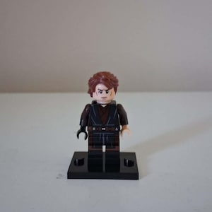 May include: A plastic minifigure with brown hair, a tan face featuring a scar over the right eye, and a dark brown and black tunic. The figure has one black hand and one tan hand, standing on a small black rectangular display base.