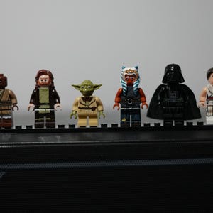May include: Six Star Wars LEGO minifigures standing in a row on a black display plate. The figures include a brown-clad character, a bearded figure in robes, a green Yoda, an orange-skinned Togruta, a black-armored Darth Vader, and a figure in white.