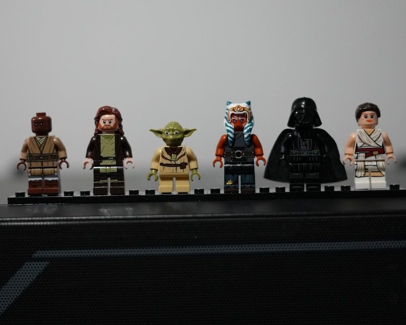 May include: Six Star Wars LEGO minifigures standing in a row on a black display plate. The figures include a brown-clad character, a bearded figure in robes, a green Yoda, an orange-skinned Togruta, a black-armored Darth Vader, and a figure in white.