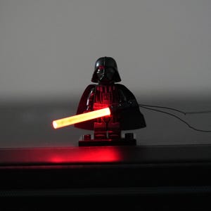 May include: A black plastic minifigure character wearing a helmet and cape, holding a glowing red cylindrical light-up accessory. The figure stands on a small black rectangular base, with a thin black cord extending from the side of the accessory.
