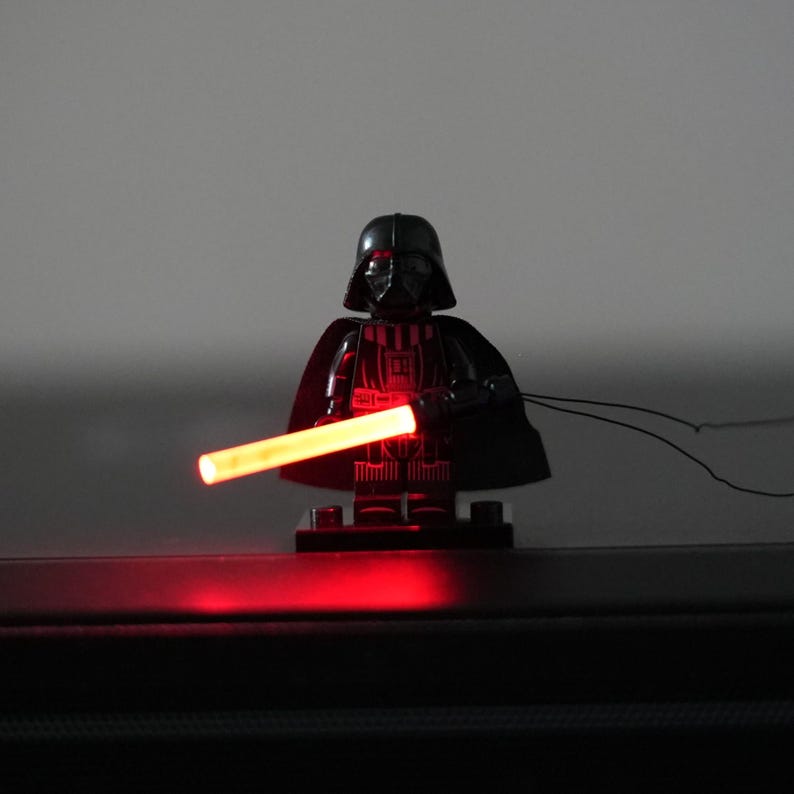 May include: A black plastic minifigure character wearing a helmet and cape, holding a glowing red cylindrical light-up accessory. The figure stands on a small black rectangular base, with a thin black cord extending from the side of the accessory.