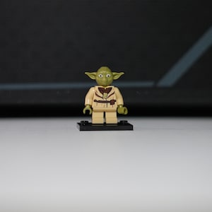 May include: A small green plastic minifigure with pointed ears, large eyes, and a tan tunic featuring a dark brown belt and collar detail. The figure stands on a small black rectangular base with two circular studs on the top surface.