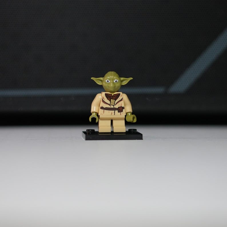 May include: A small green plastic minifigure with pointed ears, large eyes, and a tan tunic featuring a dark brown belt and collar detail. The figure stands on a small black rectangular base with two circular studs on the top surface.