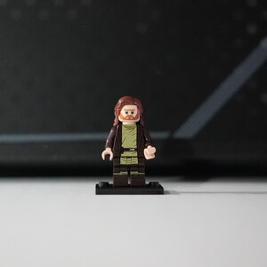 May include: A Lego minifigure featuring a male character with long brown hair and a beard. The figure wears a dark brown open jacket over a light olive green tunic with a black belt. The minifigure stands on a small black rectangular base.