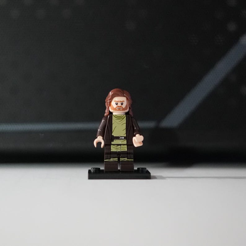 May include: A Lego minifigure featuring a male character with long brown hair and a beard. The figure wears a dark brown open jacket over a light olive green tunic with a black belt. The minifigure stands on a small black rectangular base.