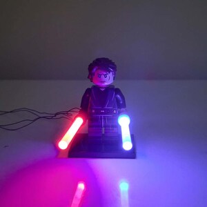 May include: A plastic minifigure character with dark brown hair and a black outfit standing on a black base. The figure holds two glowing light-up rods, one emitting a bright red light and the other emitting a bright blue light, connected by thin wires.