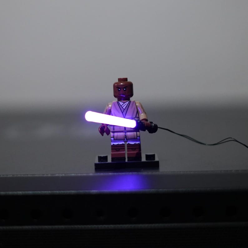 May include: A brown-skinned minifigure wearing a tan tunic and brown pants stands on a black base. The figure holds a glowing purple lightsaber accessory that is connected to a thin black wire extending to the right side of the frame.
