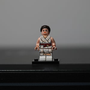 May include: A plastic minifigure with dark brown hair, a peach-colored head with printed facial features, and a white torso and leg piece featuring a draped fabric design with a dark red belt accent, standing on a small black rectangular base.