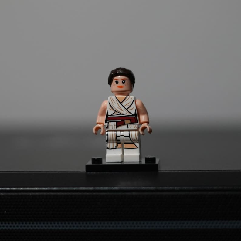 May include: A plastic minifigure with dark brown hair, a peach-colored head with printed facial features, and a white torso and leg piece featuring a draped fabric design with a dark red belt accent, standing on a small black rectangular base.